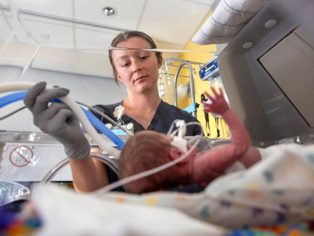A respiratory therapist wearing scrubs and gloves adjusts breathing tubes for a premature infant lying in an incubator.