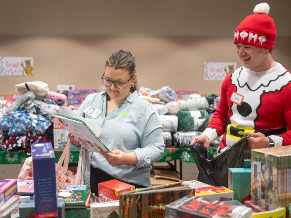 In a room full of gifts such as blankets and books, a woman looks at a children’s book. A man dressed in a seasonal “elf” outfit looks on, as he holds a bag.