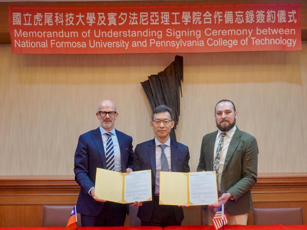 Three people sit at an official table with a flag behind them