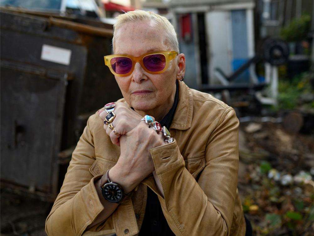Poet Jan Beatty sits outdoors in a cluttered yard or alleyway, wearing a tan jacket and orange-tinted sunglasses. She rests her hands under her chin, showing several large rings, and looks directly at the camera.