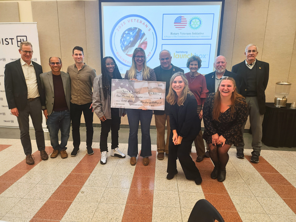 A group of 11 people stand together, one person is holding an oversized check, they all stand in front of a screen