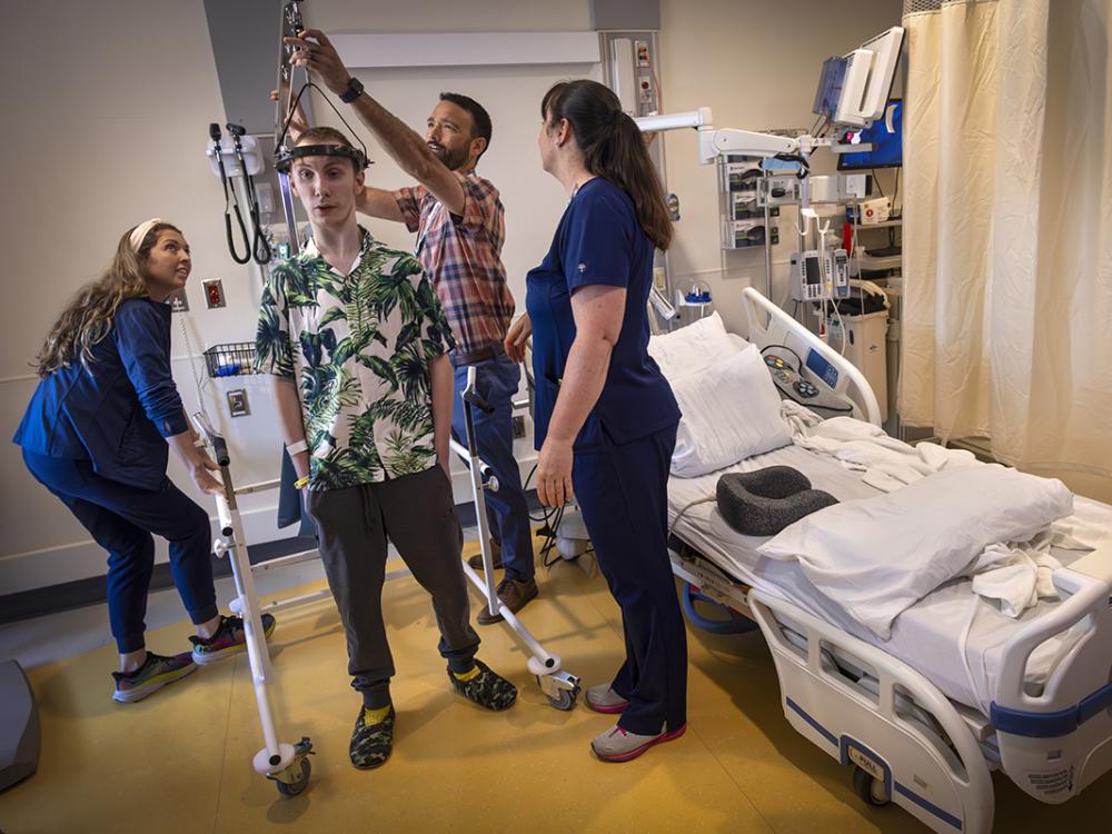 A teen boy stands wearing a halo-gravy traction device around his head. A doctor on his right adjusts the bar holding the halo up. Two nurses wearing scrubs are on the right and left of the boy and the doctor. A hospital bed and curtain are on the group’s right.