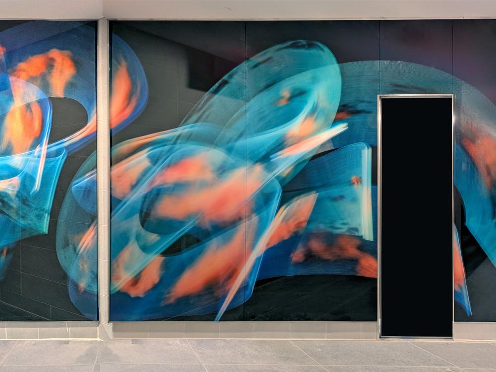 A large, abstract mural of sweeping blue and orange light forms installed along a wall inside Pittsburgh International Airport’s new terminal.