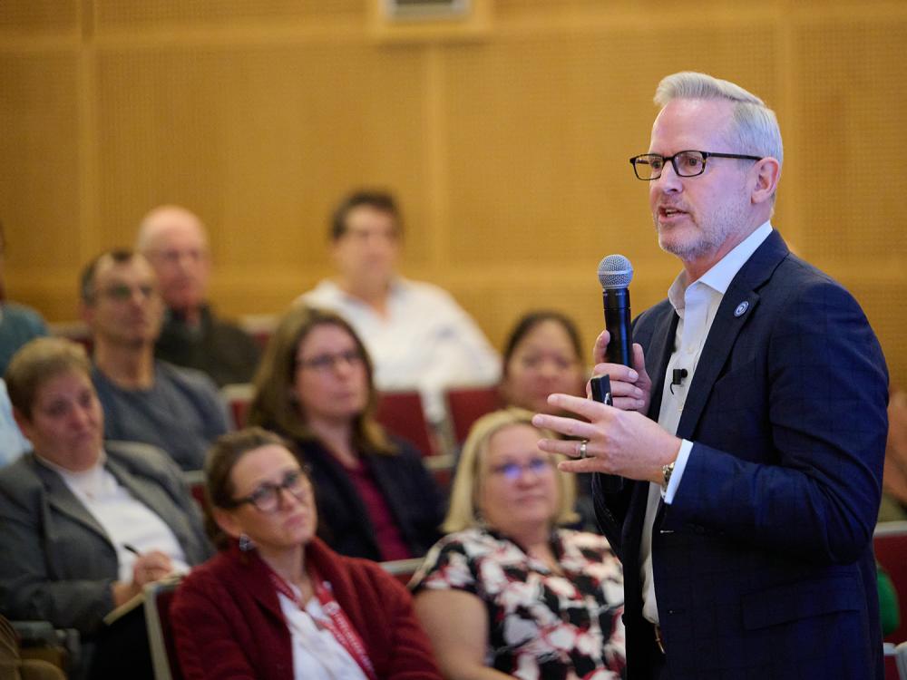Smeal College of Business Dean Corey Phelps, dressed in a blue blazer and white shirt and wearing glasses, stands in an auditorium with a microphone in his hand.