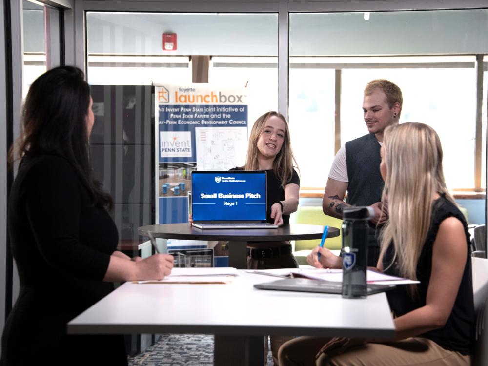Two women and a man watch another woman present her laptop, upon which is a screen that reads "Small Business Pitch: Stage 1"