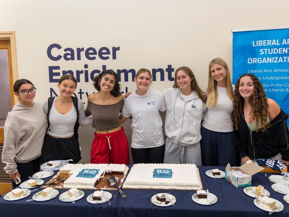 Seven members of the Liberal Arts Undergraduate Council pose with arms around each other behind a table filled with two large cakes and cake slices at LAUC’s 50th Anniversary Kickoff Celebration.