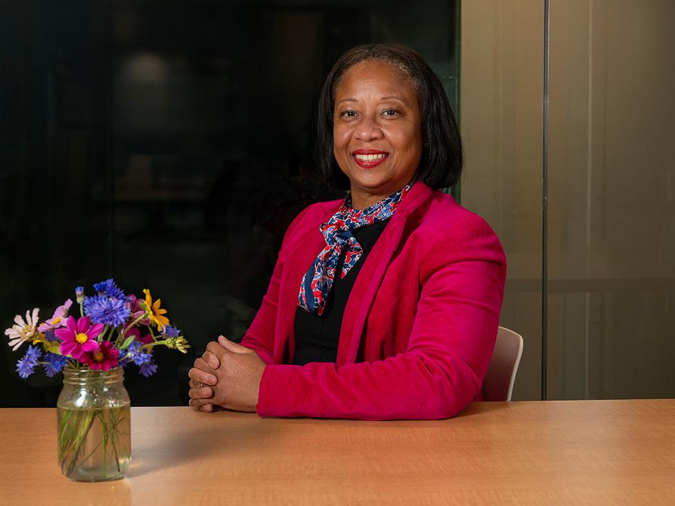 A woman sits at a table with her hands folded in front of a vase of colorful flowers. 