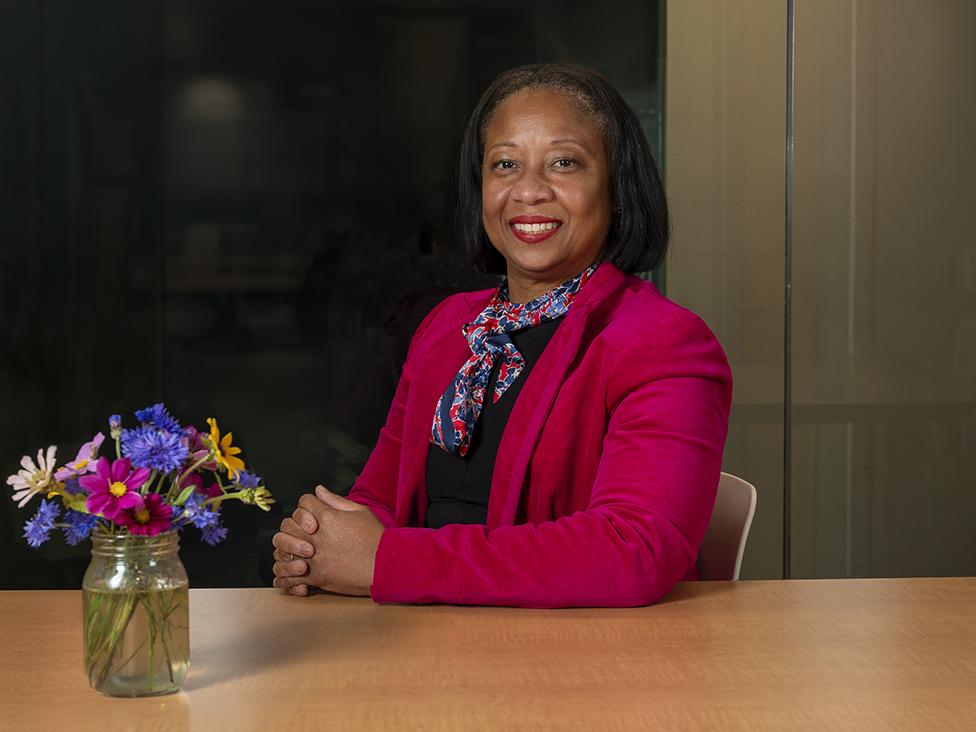 A woman sits with her hands folded on a table in front of a small bouquet of flowers.