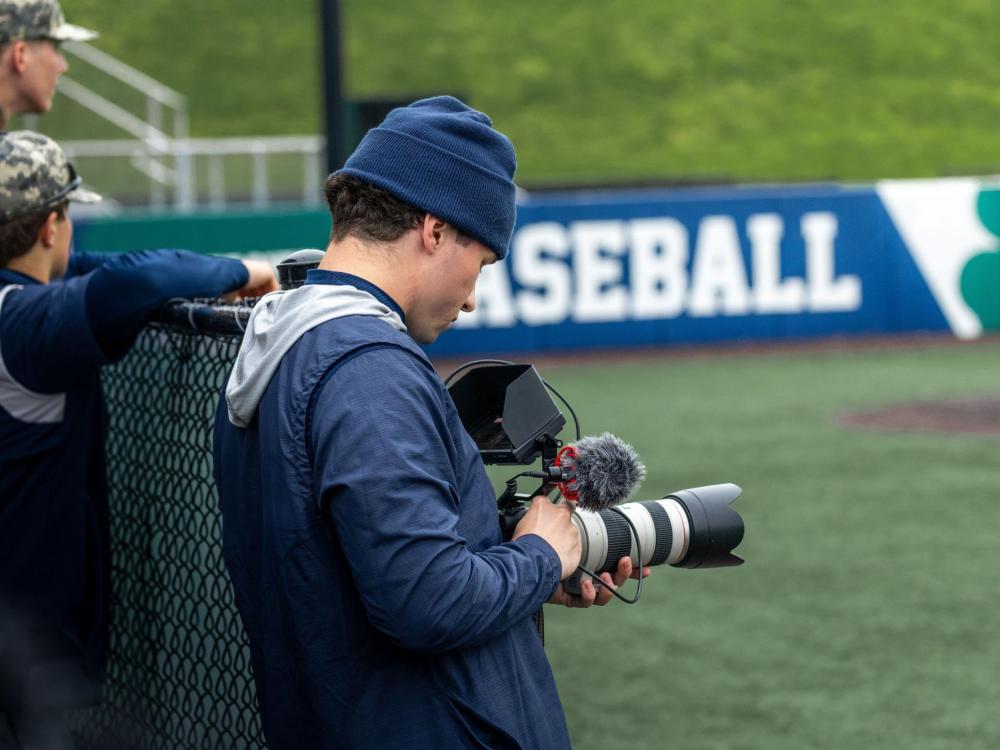 A Penn State Behrend student-athlete photographs a baseball game from the side of the field.
