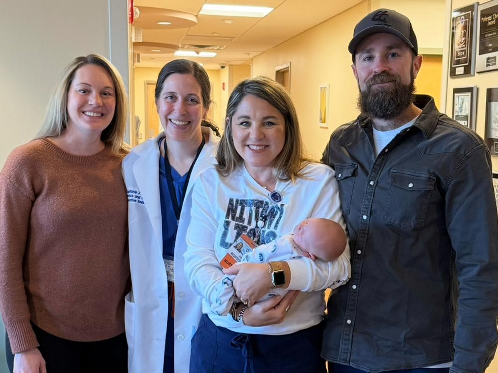 Four people, one of whom is holding a baby, smile as they pose for a photo in the hallway of a doctor’s practice.