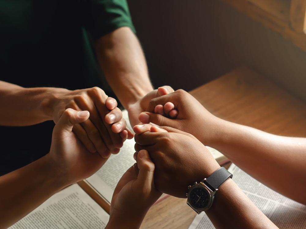 Group of people holding hands in a circle over an open book on a table, symbolizing unity and support.