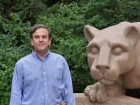 Mark Roberts in front of the Nittany Shrine in a light blue button-down shirt, grey pants, with short brown hair smiling.