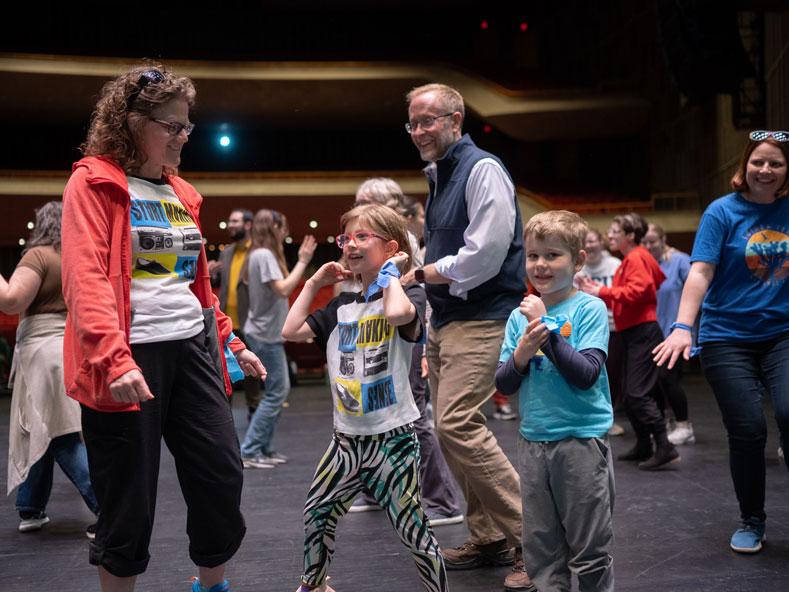 A family of two parents and two children dance on a stage.