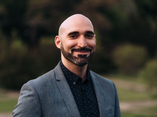 A bearded man in a blazer smiles at the camera in an outdoor setting