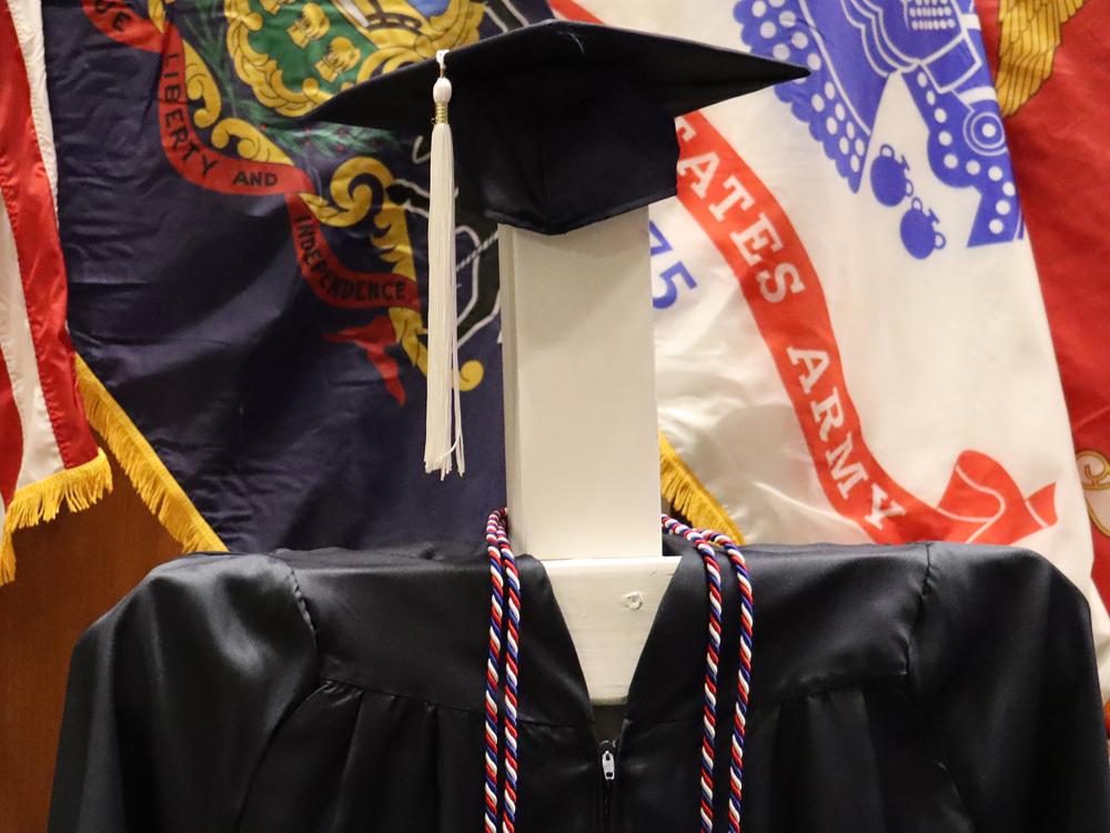 A blue graduation cap and gown with red, white, and blue honor cords in front of a series of U.S. military flags.