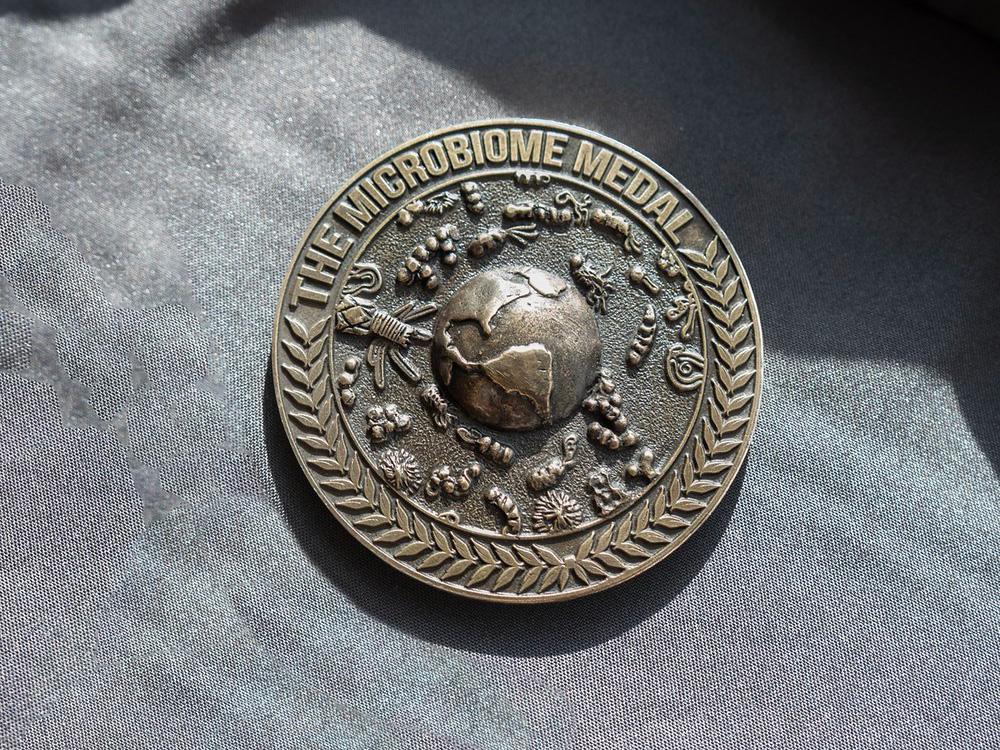 A bronze medallion with the words "The Microbiome Medal" above a globe surrounded by microscopic life