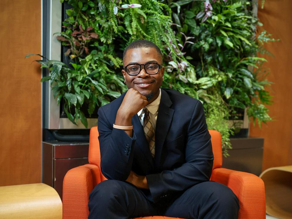 Barron Owusu wears a suit while sitting on a bright orange chair in front of plants.