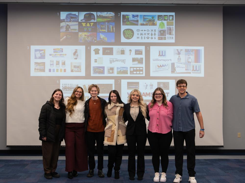 Seven students stand in front of a projection screen for a photo