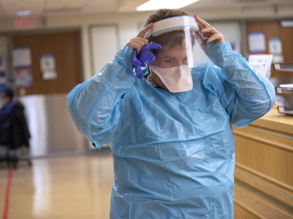 A nurse dressed in a plastic gown, mask and face shield adjusts the face shield as she walks through a hospital hallway.