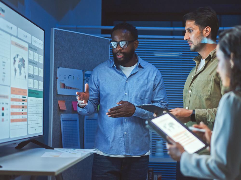 Two men and a woman, casually dressed stand in front of a monitor displaying graphs, maps and statistics