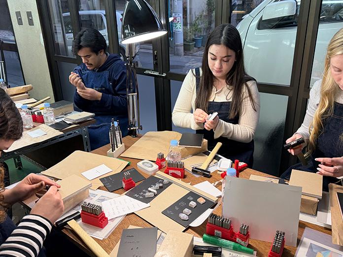 Four college students sit around table with art supplies and tools making art