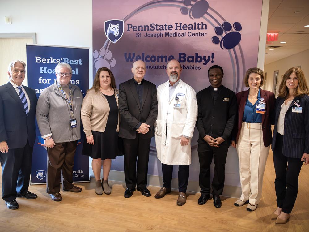 A group of eight people pose for a photo at the Breidegam Family Birthing Center at St. Joseph Medical Center. They stand in front of two banners. The one behind them reads “Penn State Health St. Joseph Medical Center” and “Welcome Baby!”