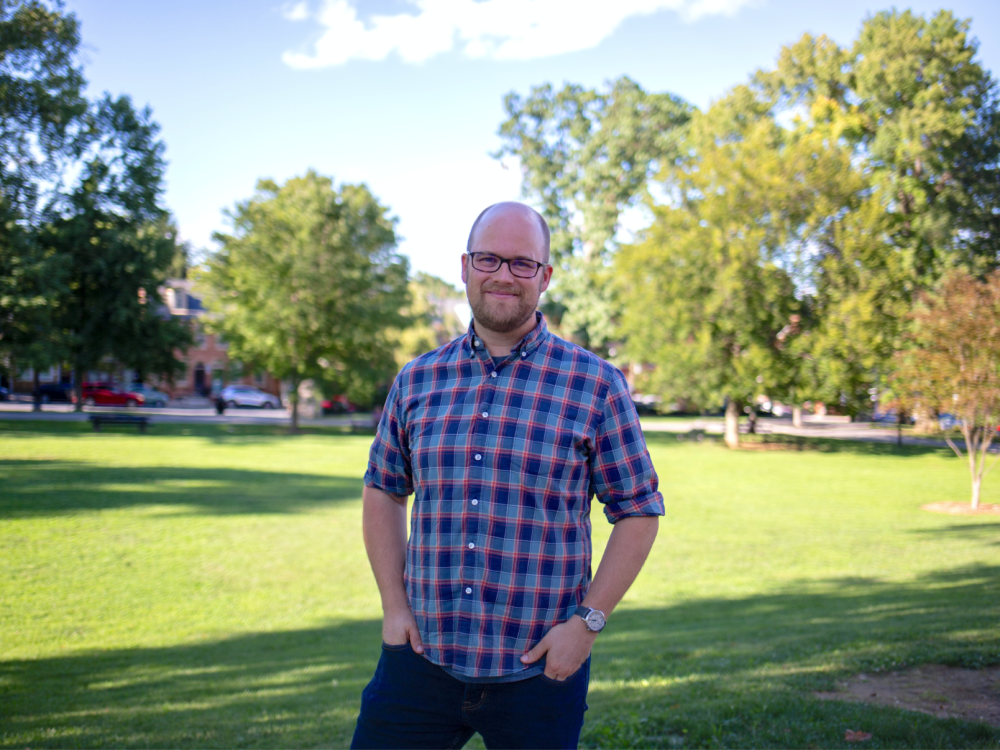 A man stands outside in a plaid short-sleeved shirt with a lawn and trees behind him. 