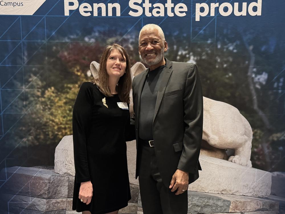 A man and woman stand in front of a Penn State photo background.