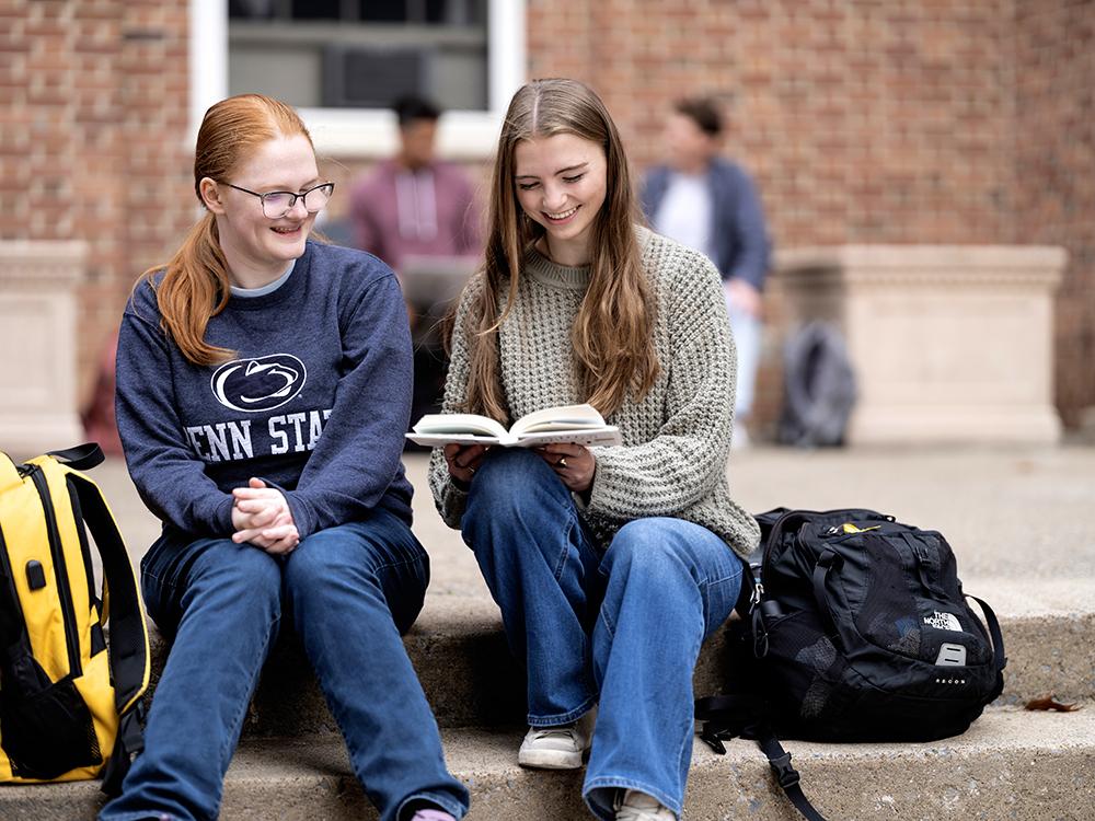 Two girls seated on a set of outdoor concrete stairs with the girl on the right reading a book while the other one looks on