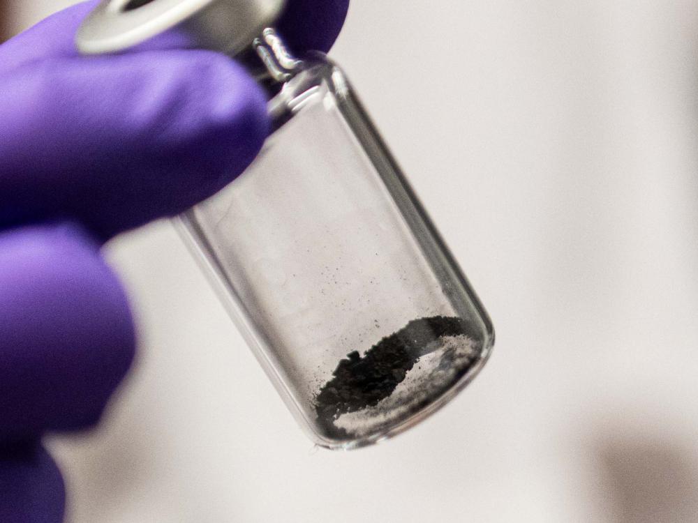 A gloved hand holds a vial with dust