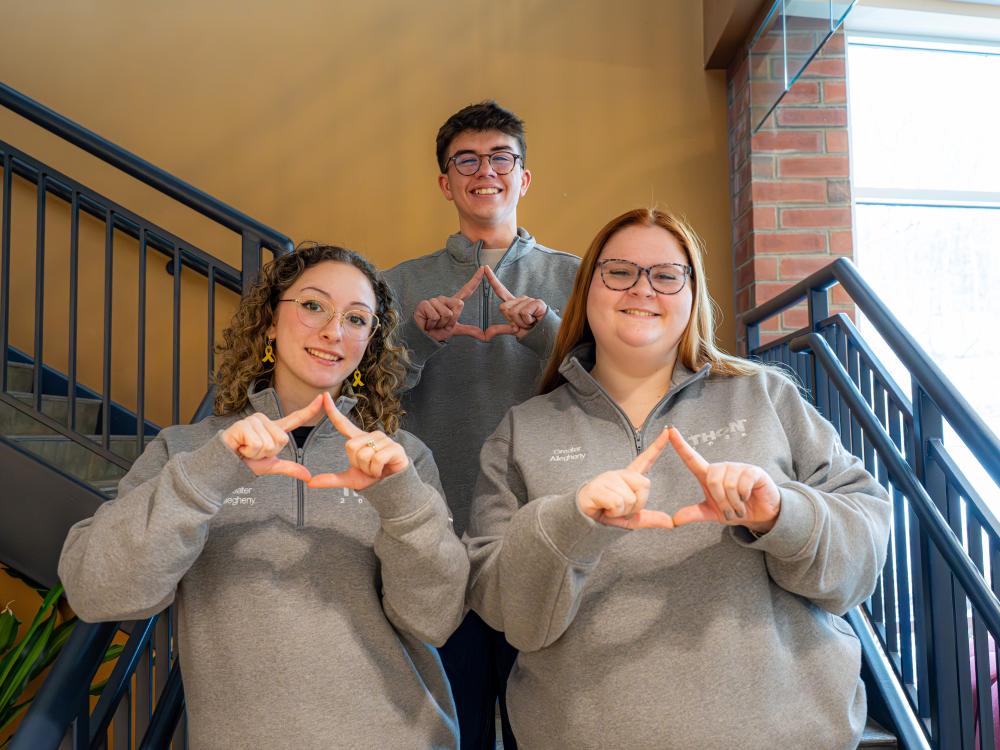 Three Greater Allegheny Benefiting THON student leaders stand on a staircase, each wearing a gray quarter‑zip with organization text on the chest. They hold their hands in the THON diamond gesture, with two students in front and one standing centered behind them. The setting includes metal railings, warm-colored walls, and natural light coming from a nearby window.