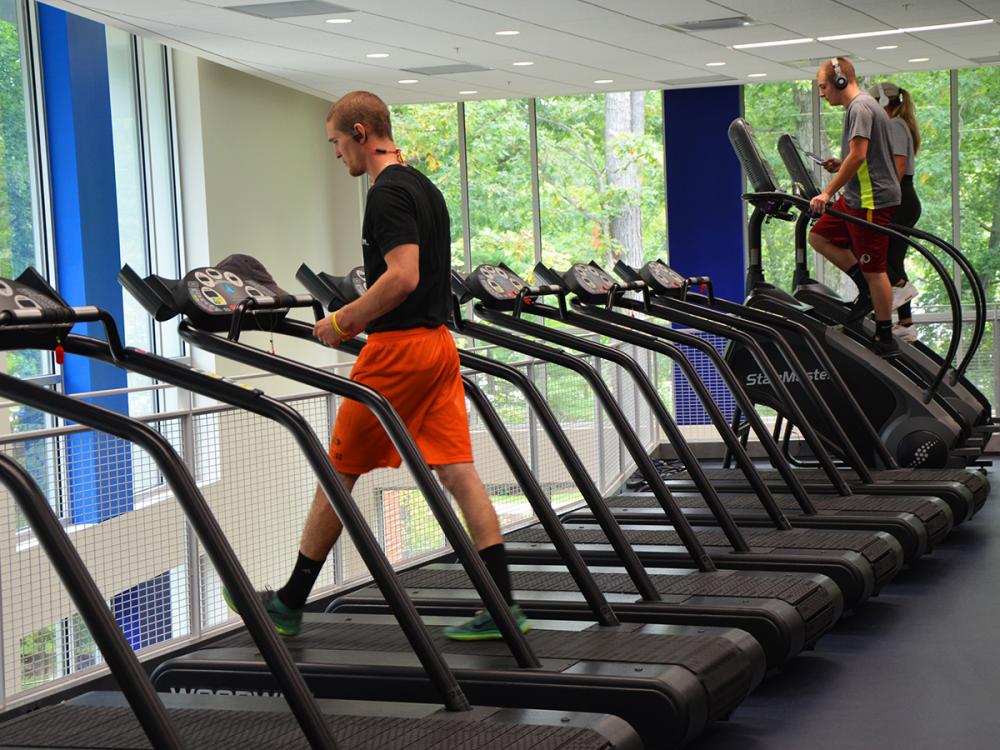 Students exercising on treadmills and stair machines inside a bright fitness center with large windows overlooking trees.