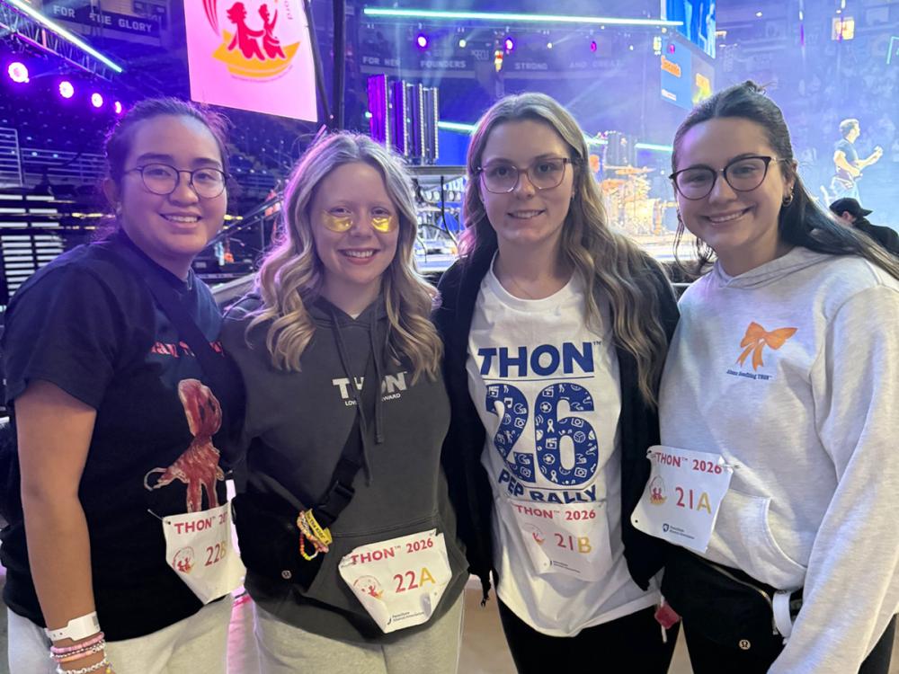 Four students pose together at a THON event, smiling in front of a brightly lit stage. They are wearing casual attire and participant bibs labeled with THON 2026 sections.