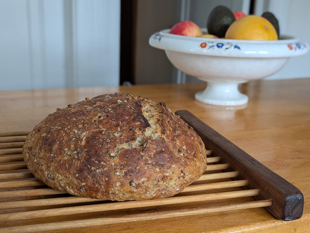 A loaf of whole grain bread sits on a cooling rack on a kitchen island. In the background is a bowl of fruit including grapefruit, avocado, and apple.