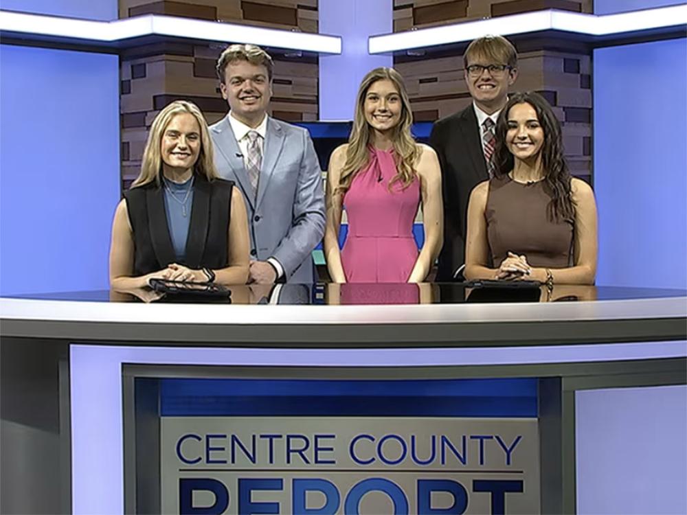 Five student standing behind an anchor desk in a TV studio