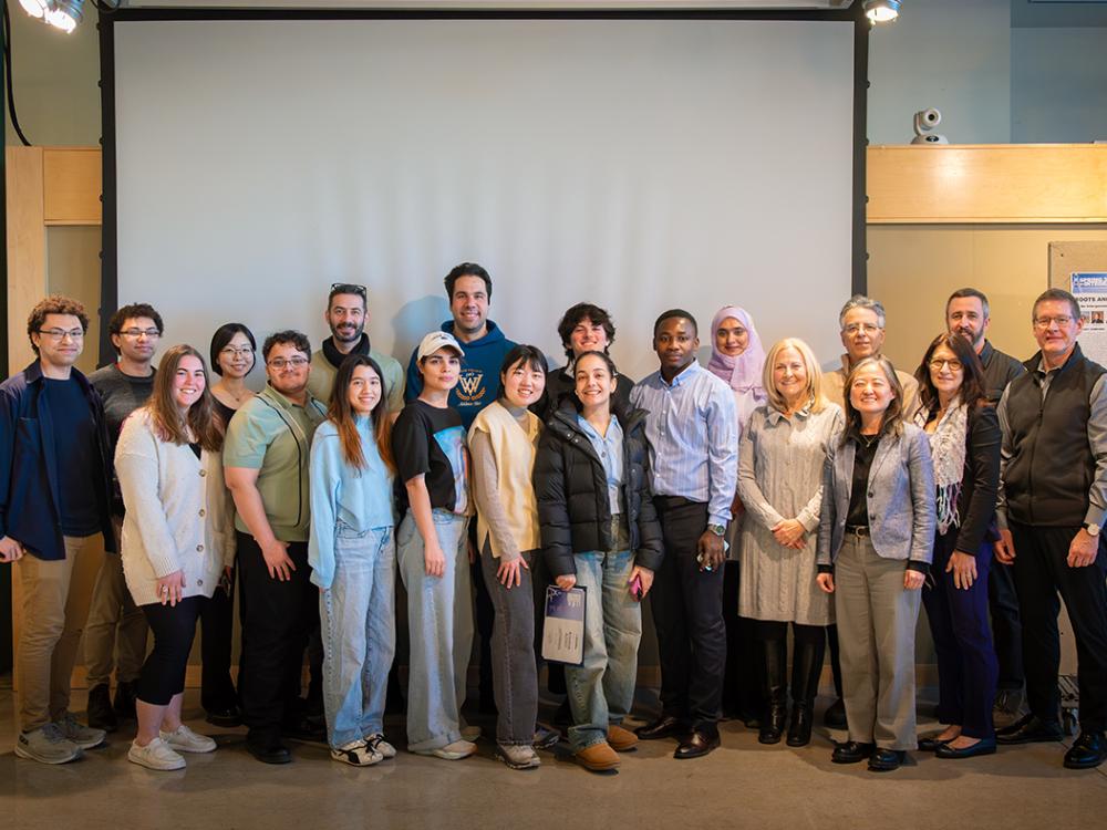 A large group of people stands together in front of a blank projection screen in a well-lit indoor space. 