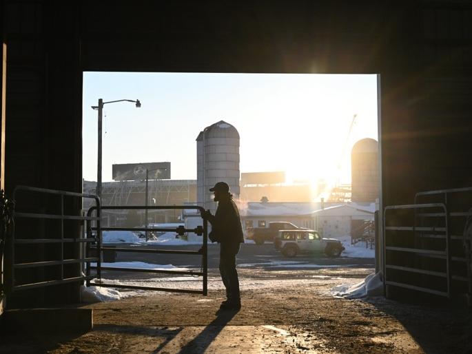 An individual stands in the shadows in front of an open barn door with a light-filled sky in the background