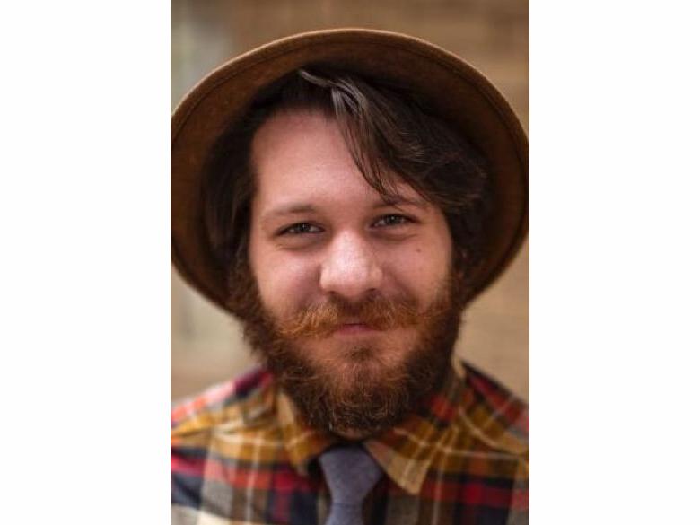 A headshot Jacob Hannah smiling with a brown beard and mustache and a plaid shirt and brown derby hat.