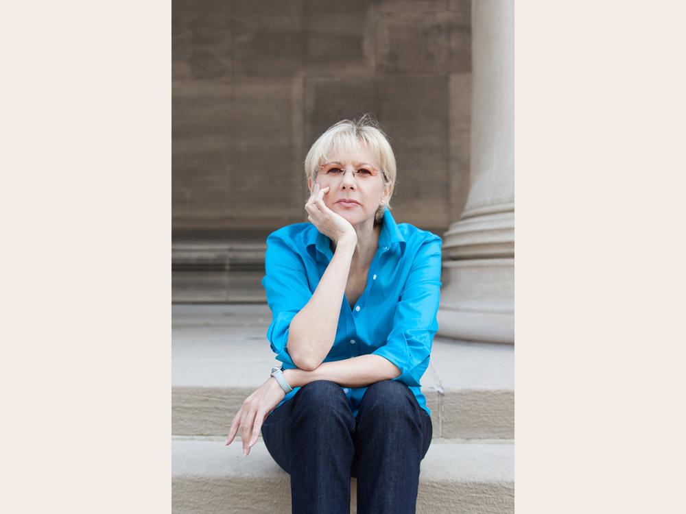 Lynn Emanuel wearing a light blue blouse with chin resting on hand while sitting on steps