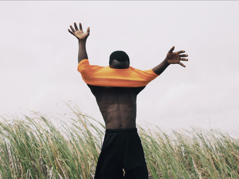 Photograph of a man standing in front of tall grass with his arms outstretched and an orange shirt covering his face