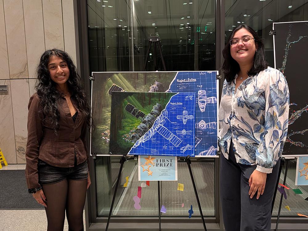 Two Penn State students standing with their artwork in between them on an easel. The artwork is a painting of a satellite, where on the left side of the painting the satellite is withered on a forest floor, and on the right side it is a drafted blueprint.   