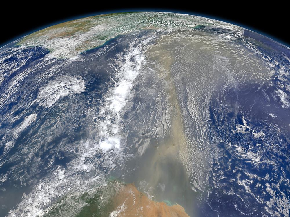 Satellite view of Earth showing the Sahara Desert in the foreground, with a vast plume of dust extending over the Atlantic Ocean under a cloudy sky.