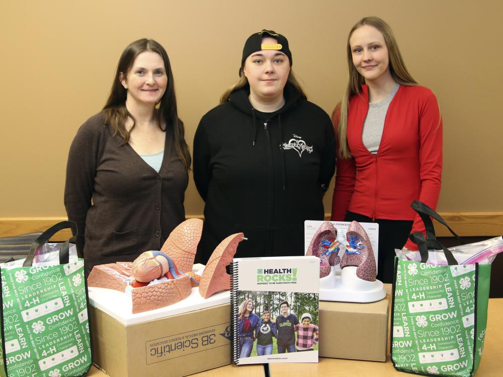Tiffany Morris, Kenzie Ulan and Raelin Dennis pose for a photo standing behind a table with Health Rocks books and models of human organs.