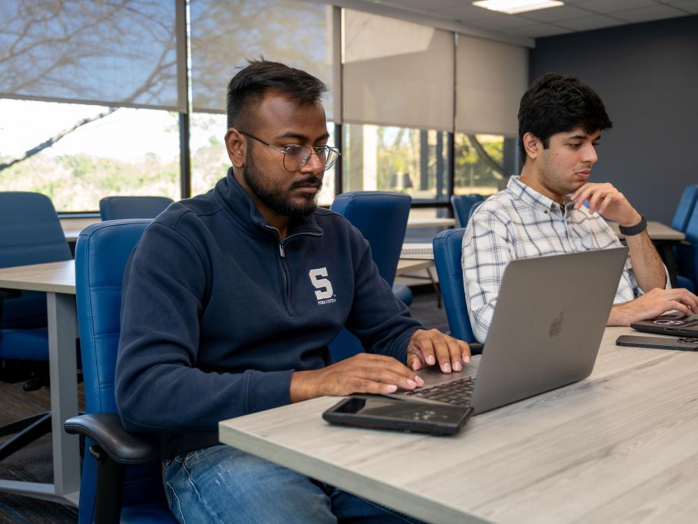 Two students sit at a table working on their laptops.