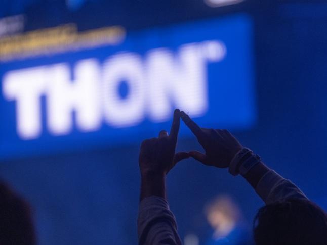 A person in the shadow raises their hands in the shape of a diamond in the foreground of a lit "THON" sign