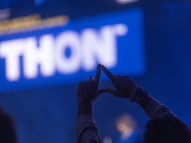 A person in the shadow raises their hands in the shape of a diamond in the foreground of a lit "THON" sign