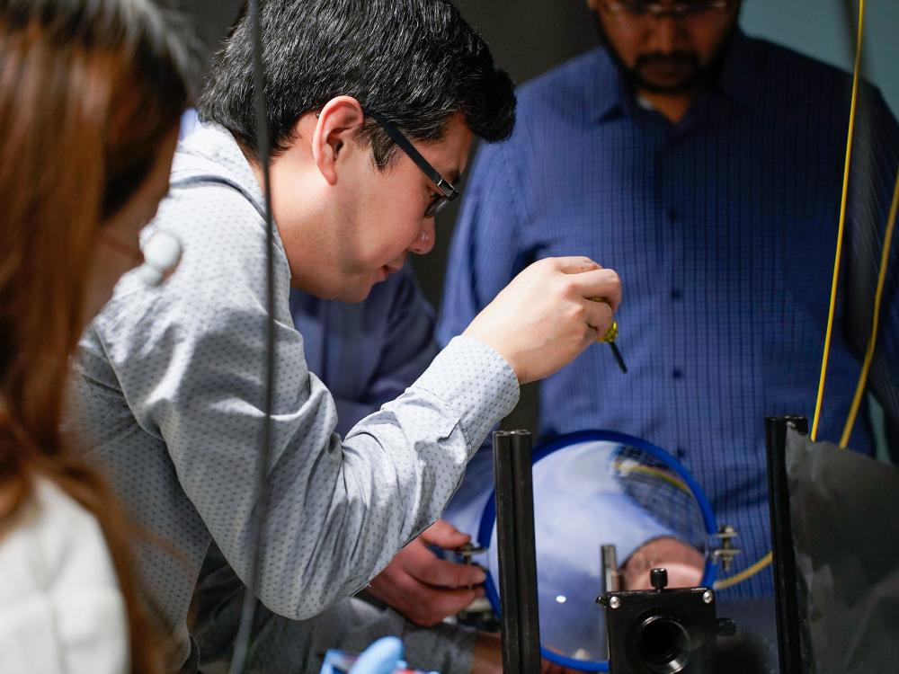 A photo of Xingjie Ni leaned in with a screwdriver, working on a machine consisting of optical lenses and mirrors.