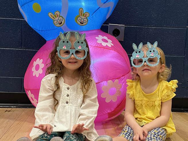 Two young girls wearing bunny glasses and sitting on the floor. 