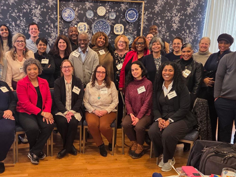 A large group of symposium attendees pose together for a group photo in a room with patterned wallpaper and a decorative display of plates on the wall