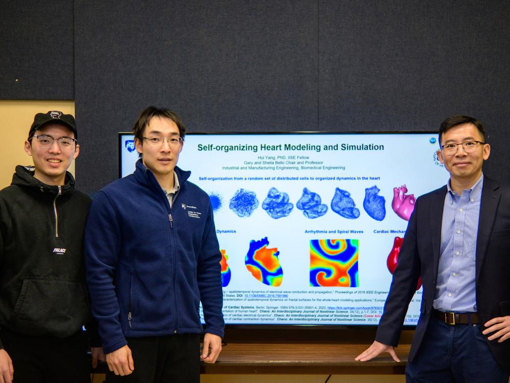 Three men pose for a photo in front of a television that reads "Self-organizing Heart Modeling and Simulation" with a series of graphics of graphics showing the heart in different phases of self-organization, with electrical maps of a heart beneath it. 
